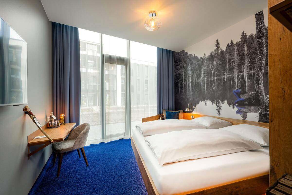 Habitación de hotel con una cama grande, un escritorio con una silla, un televisor y un mural con una escena de bosque.