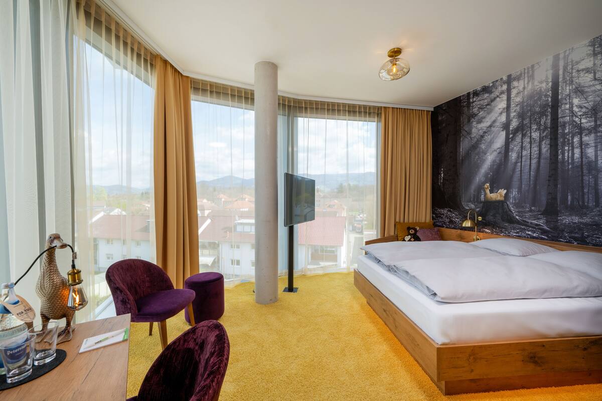 Habitación de hotel con una cama grande, un escritorio con lámpara, una silla y vistas a la ciudad a través de amplios ventanales.