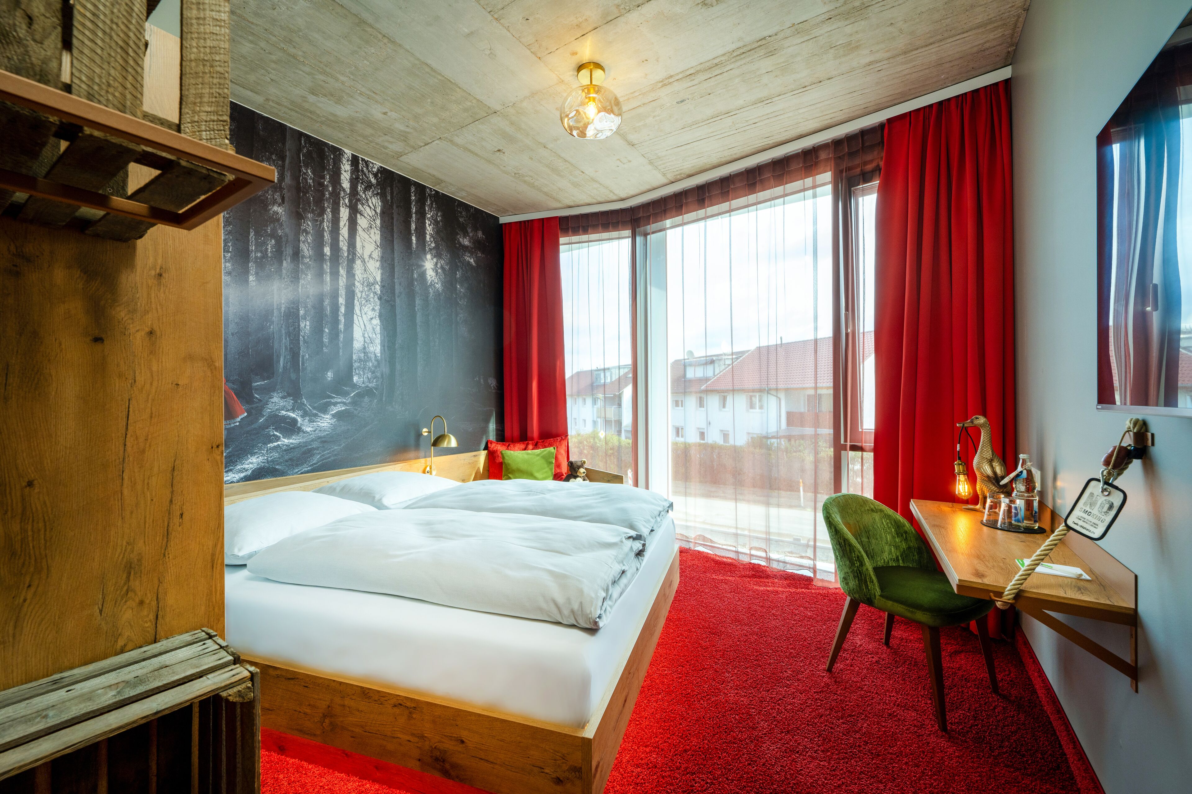 A hotel room with a large bed, a desk with a chair, a red carpet, and a large window with red curtains.