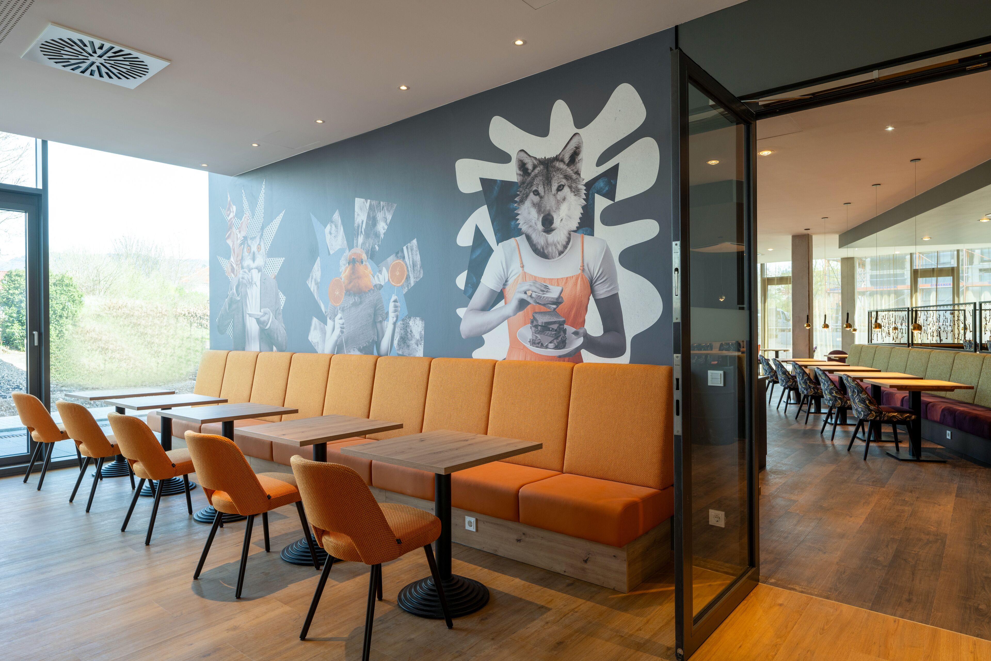 Een modern restaurant met oranje zitplaatsen, houten tafels en een muurschildering.