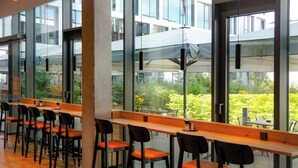 Un área de estar interior moderna con largas mesas de madera y sillas negras, grandes ventanales que ofrecen vistas a zonas verdes y el exterior de un edificio.