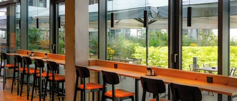 A modern indoor seating area with long wooden tables and black chairs, large windows offering views of greenery and a building exterior.