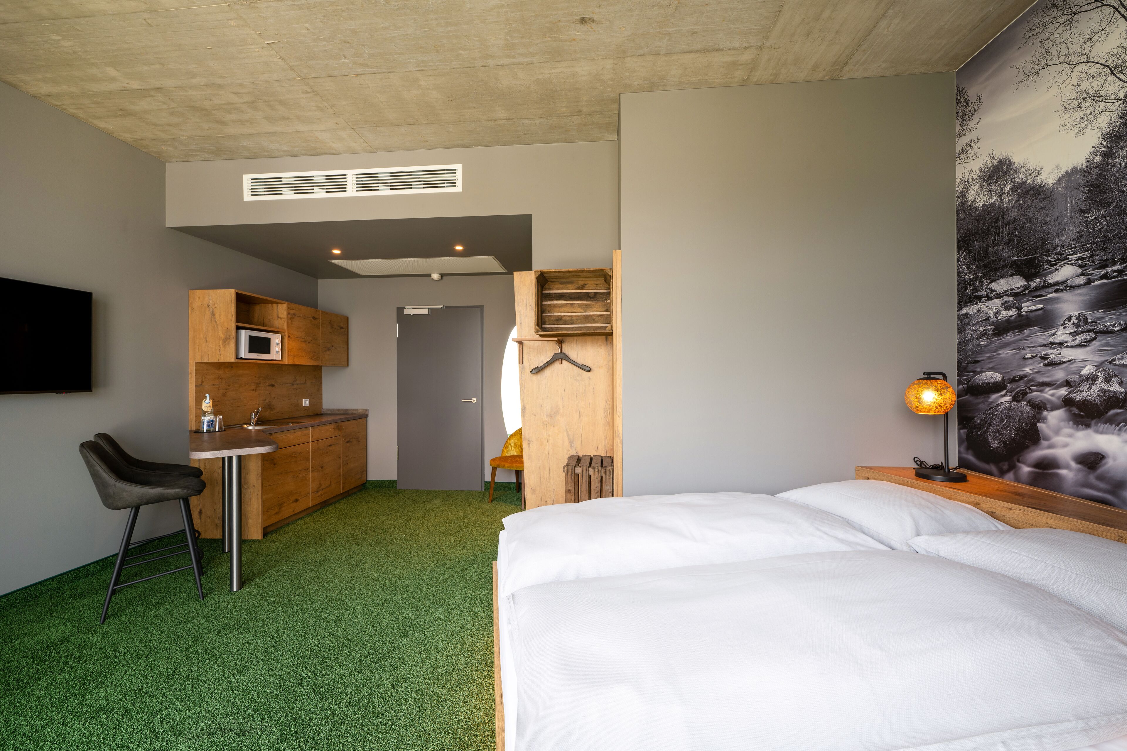A hotel room with a bed, a desk with a chair, a microwave, a small kitchenette, and a painting of a river scene.