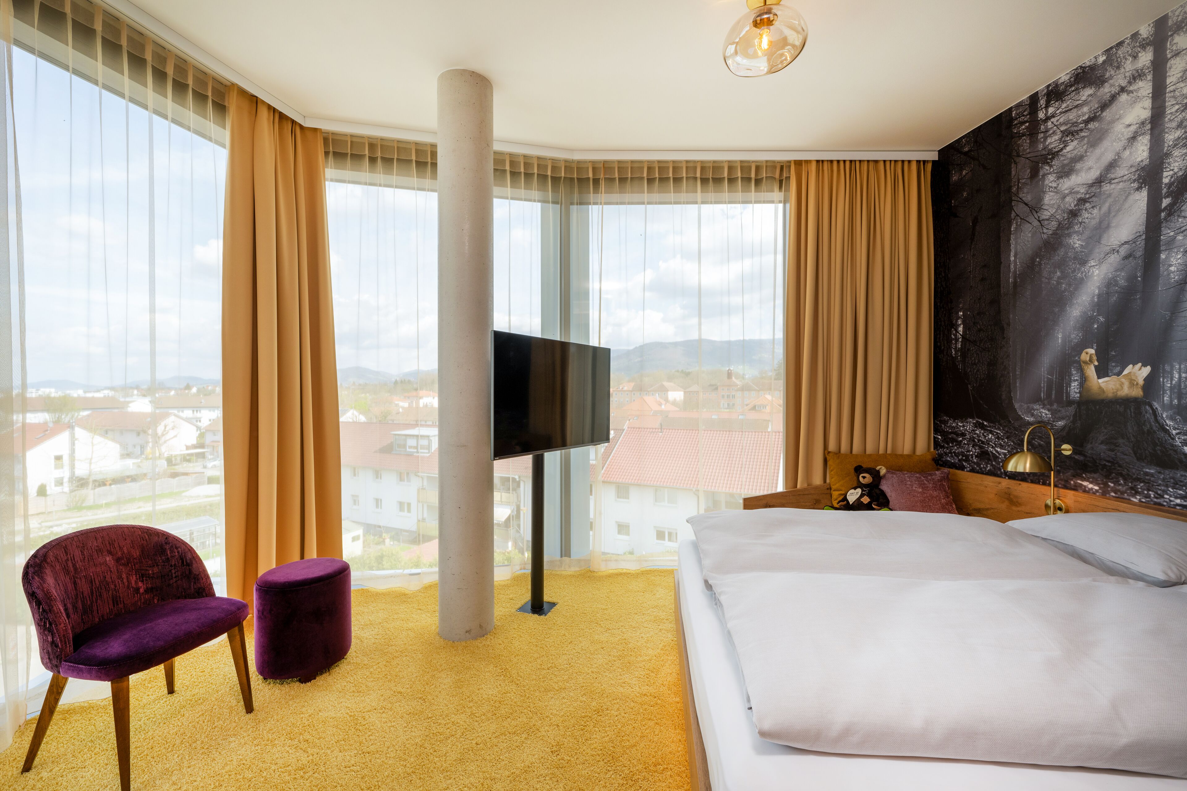 Una habitación de hotel con una cama, una silla, un televisor y un ventanal con vistas a edificios.