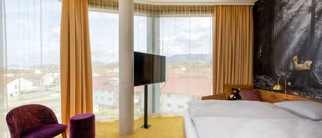 A hotel room with a bed, a chair, a television, and a large window with a view of buildings.