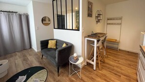 Un espai habitable compacte amb un sofà gris, una taula rodona petita, una zona de menjador amb taula i cadires blanques, i un paviment de fusta.