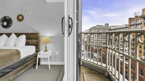 Een hotelkamer met een bed, een klein wit tafeltje met een lamp en uitzicht op de stad vanaf een balkon met een metalen hekwerk.