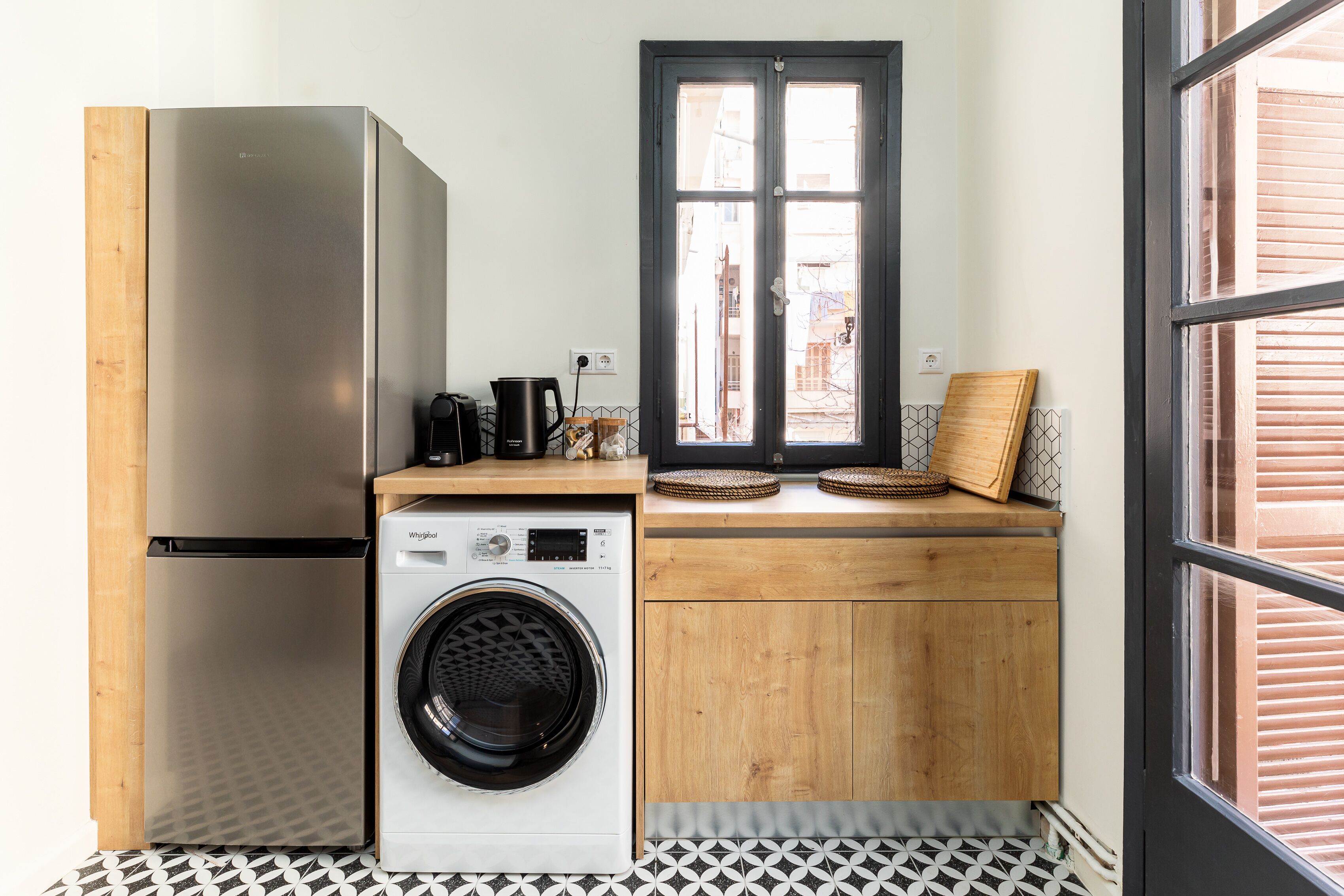 Une cuisine compacte équipée d’un réfrigérateur en acier inoxydable, d’un lave-linge, de meubles en bois et d’une fenêtre donnant sur des immeubles.