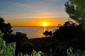 Un coucher de soleil sur l’océan, avec un promontoire rocheux au premier plan et une végétation luxuriante.
