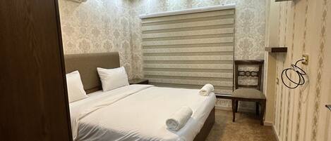 A bedroom with a bed, a nightstand, a chair, a wooden ceiling feature, and patterned wallpaper.