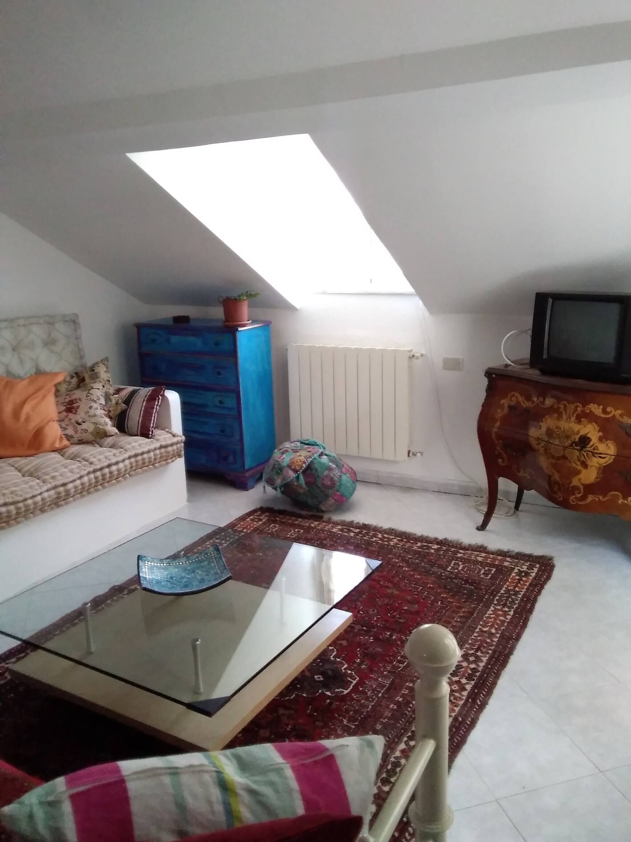 An attic room with a sofa, a glass coffee table, a television, a radiator, and a patterned rug.