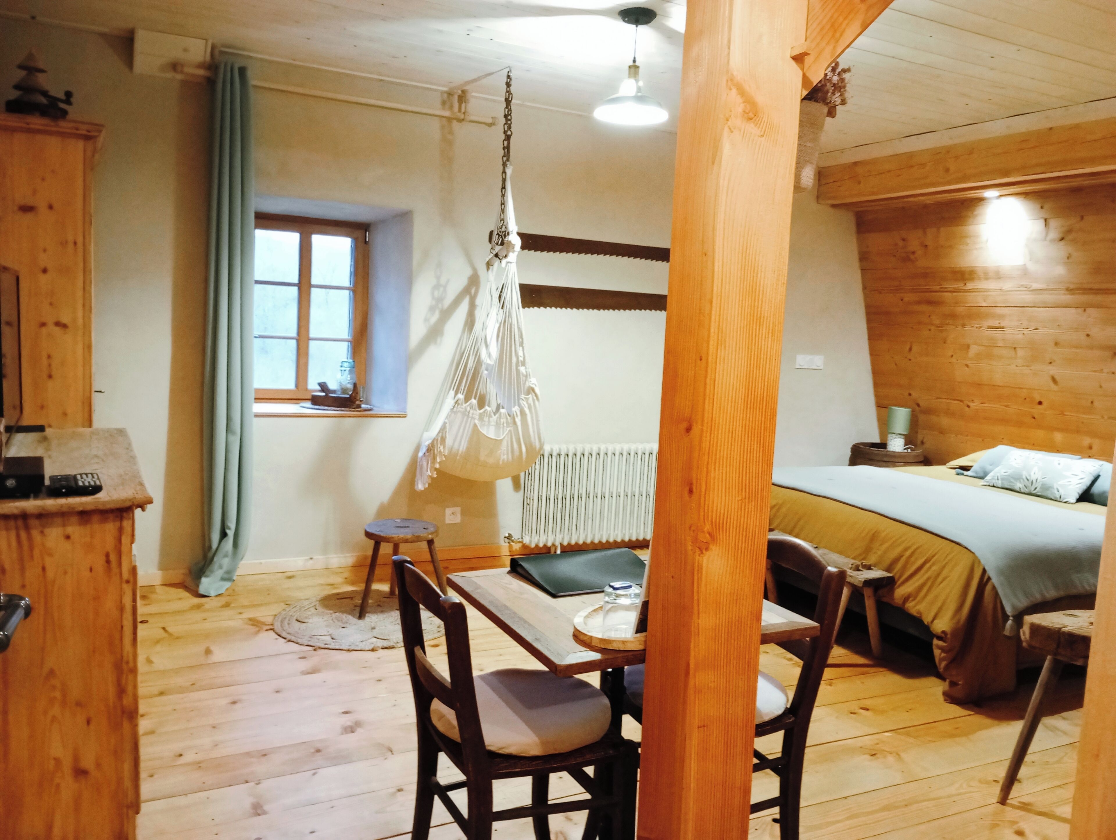 A cozy room with a wooden floor, a bed with a green blanket, a hanging chair, a small table, and a window with curtains.