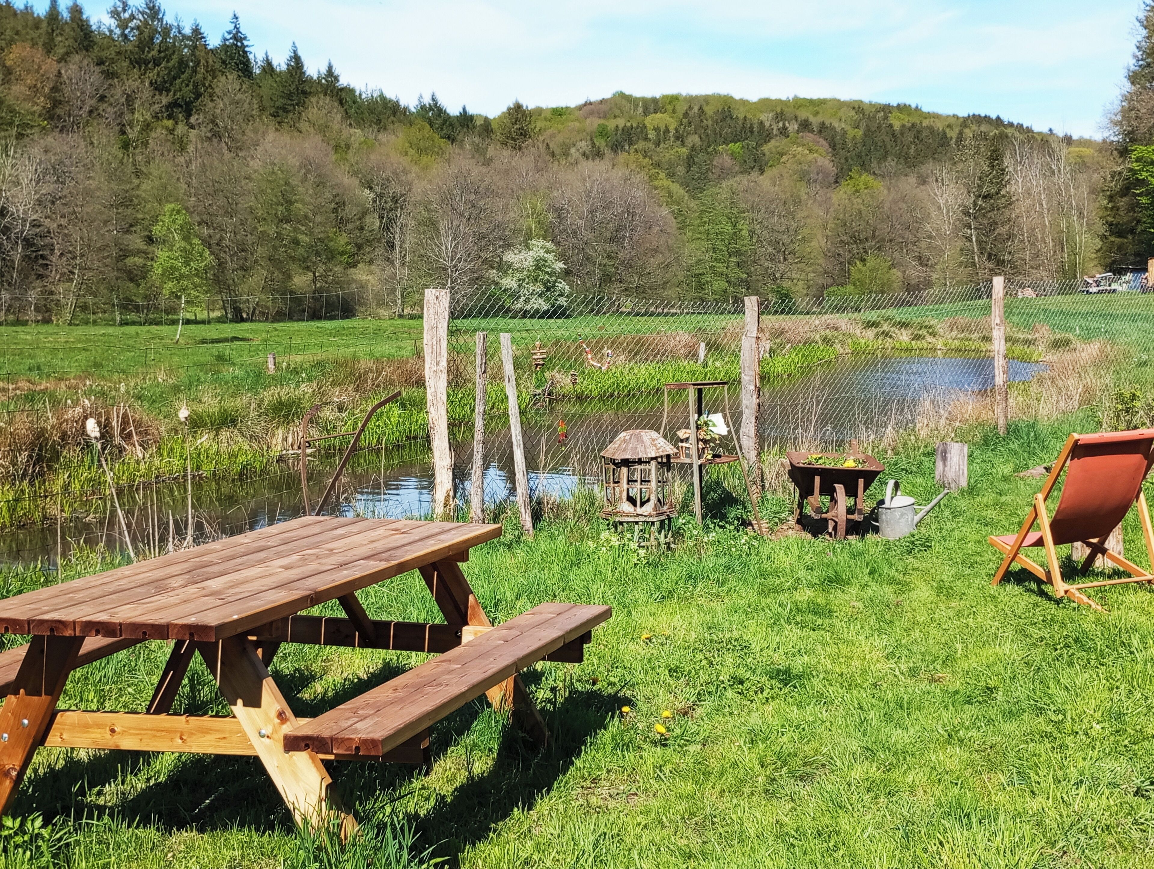 Une table de pique-nique et des chaises sont installées dans un espace herbeux, avec un étang et une clôture en arrière-plan.