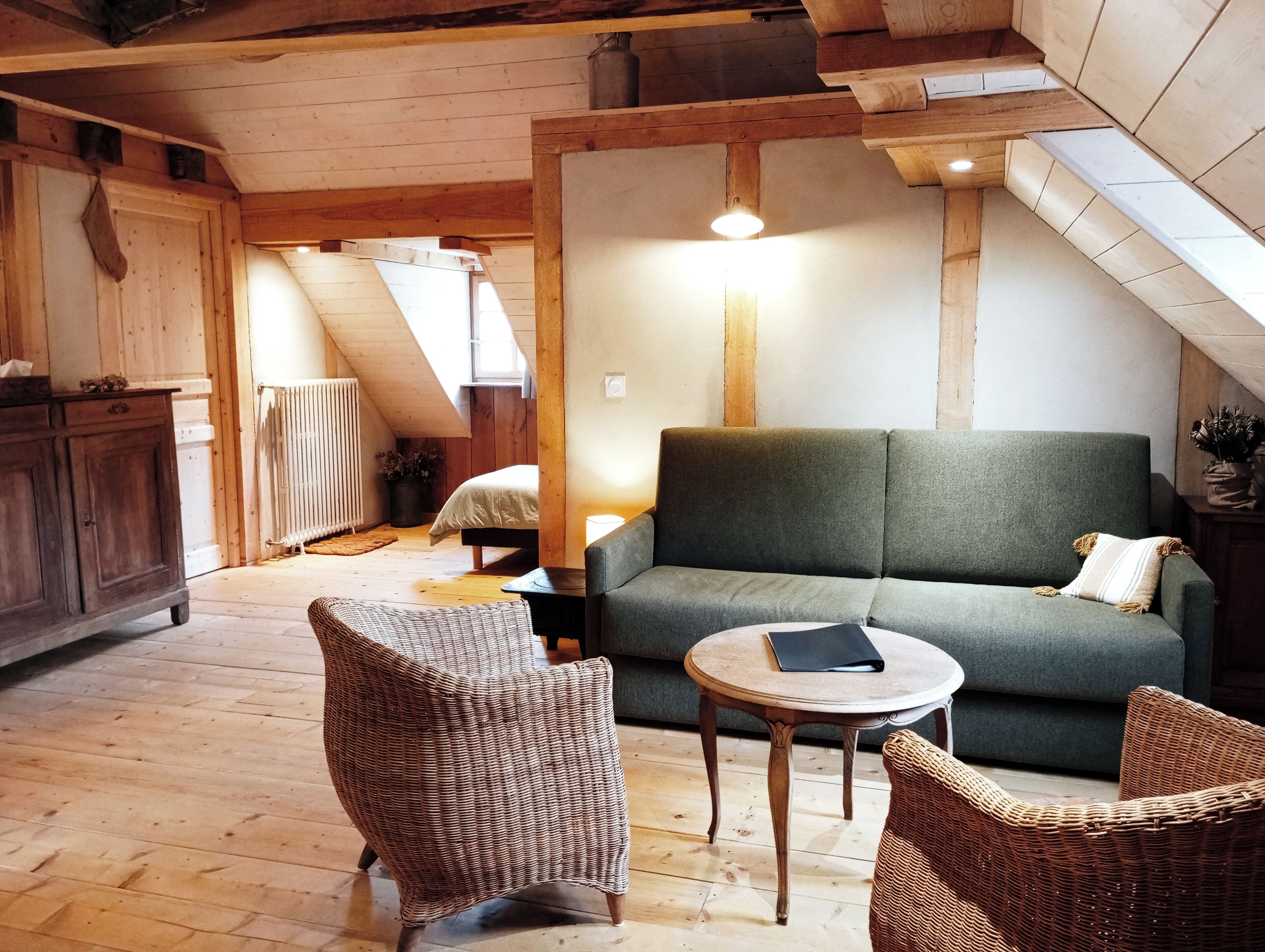 Una habitación acogedora estilo loft con un sofá verde, sillones de mimbre y una pequeña mesa redonda. Se aprecia un radiador al fondo, y la habitación tiene vigas de madera y un techo inclinado.
