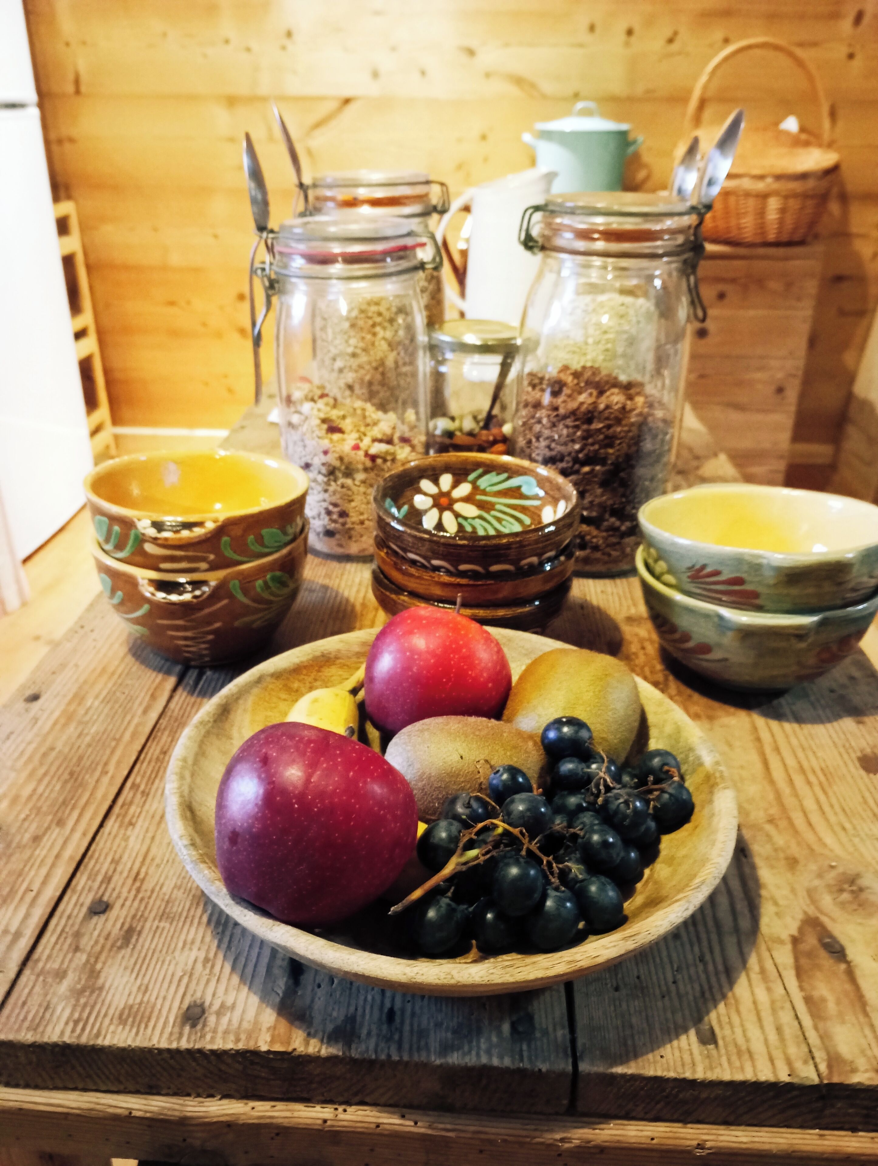 Una mesa de madera con un bol de frutas, incluyendo manzanas y uvas, y varios frascos de vidrio con granos y semillas.