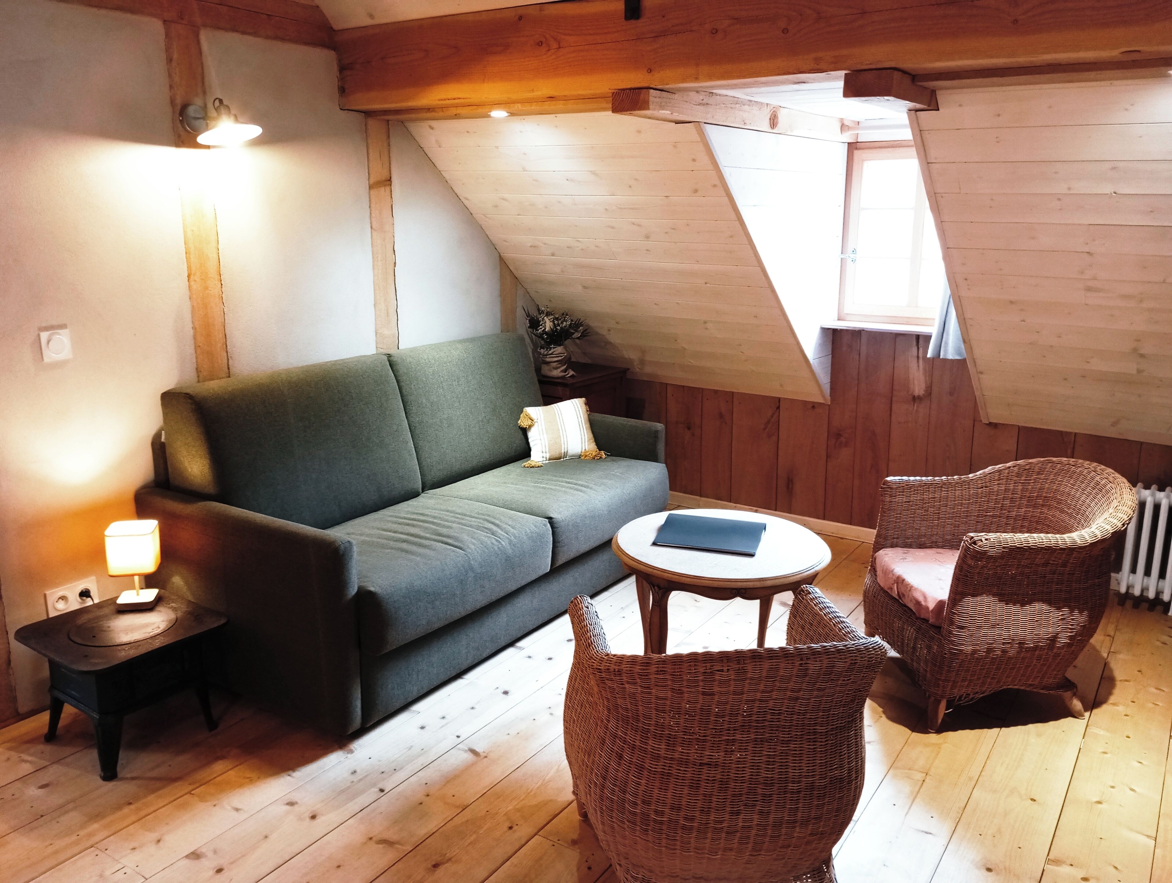 Une pièce mansardée chaleureuse, avec un canapé vert, des fauteuils en osier, une petite table et une lampe.