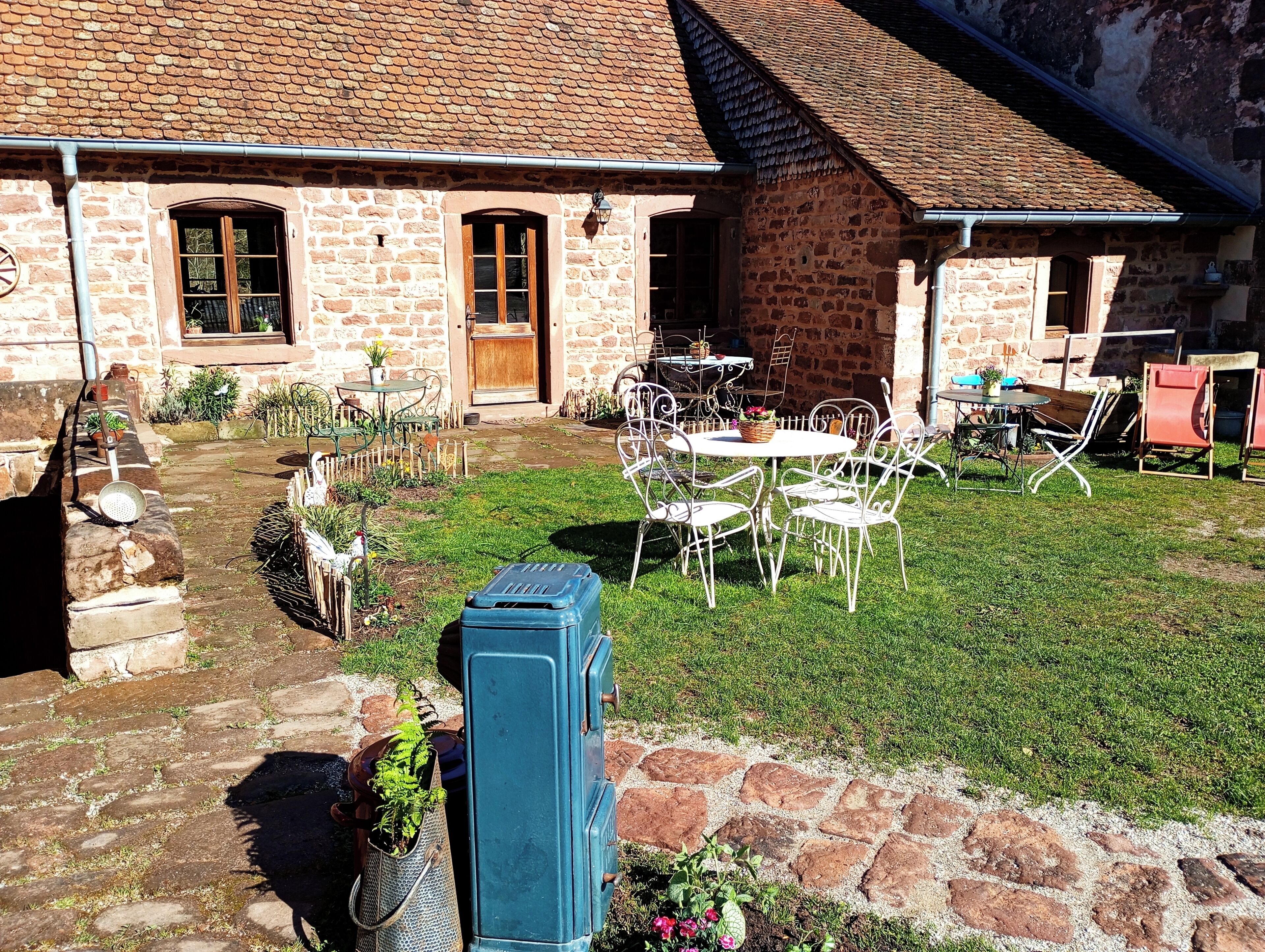 A stone building with a brick roof, outdoor seating area with metal chairs and tables, and a garden with plants and a blue storage unit.