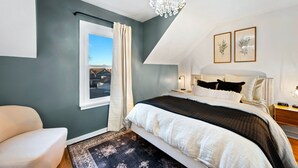 A bedroom with a large bed, a chandelier, a window with curtains, and two framed pictures on the wall.