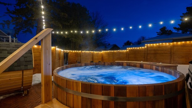 A wooden hot tub with a wooden deck and a wooden fence, surrounded by string lights.