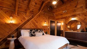 A bedroom with a wooden ceiling, a bed with white bedding, a dresser, and a round mirror.