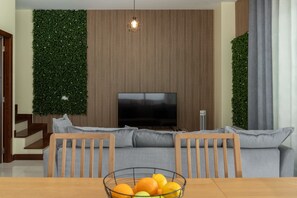 A modern living room with a flat-screen TV, a wooden dining table with a fruit bowl, and a sofa with grey cushions. - Vacation Home in Porto Santo, Villa Girassol (Porto Santo)