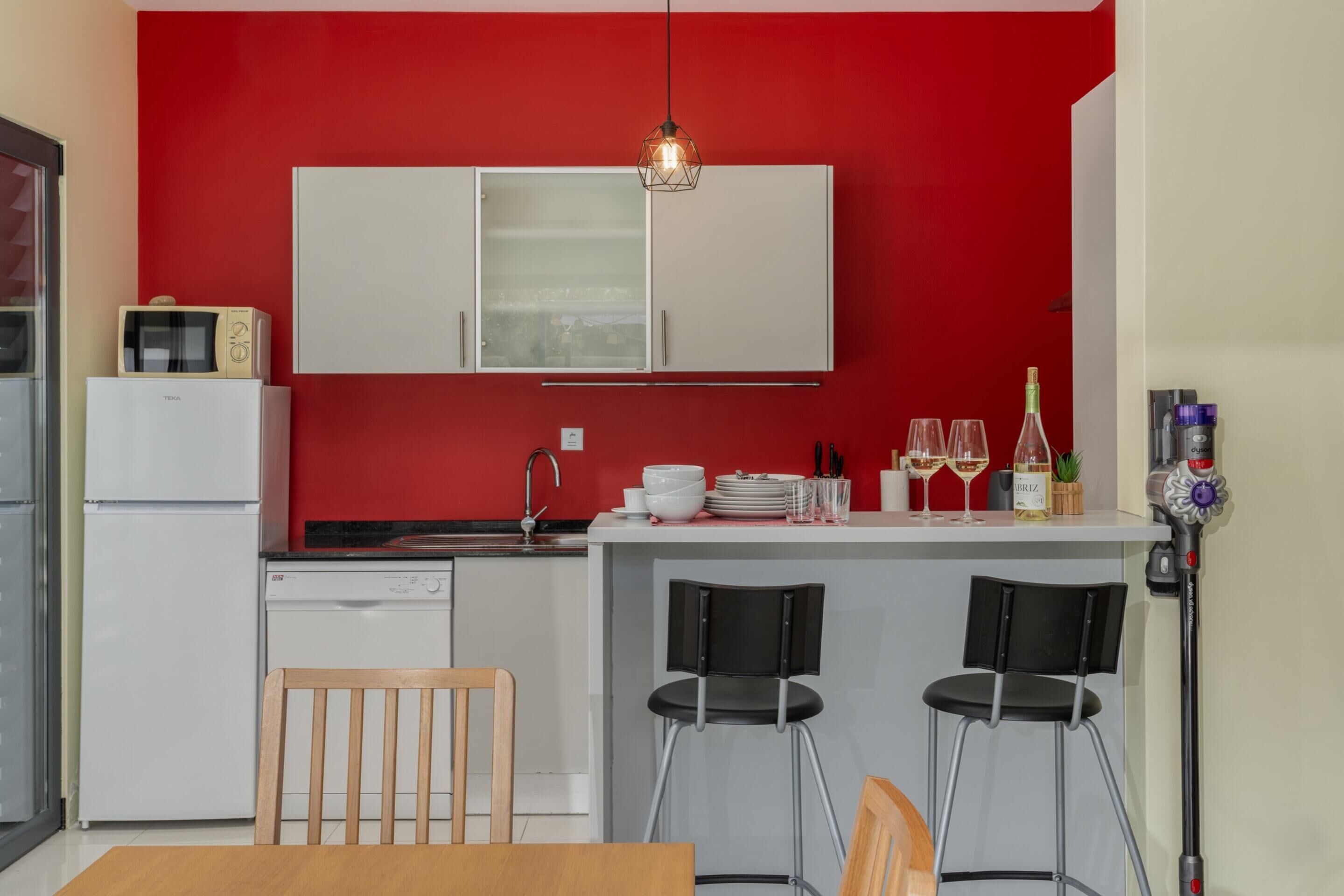 Cocina compacta con pared roja, gabinetes blancos, microondas integrado y una barra con dos taburetes.
