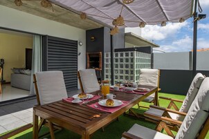 An outdoor dining area with a wooden table, white cushioned chairs, and a covered seating area with a view of the cityscape.