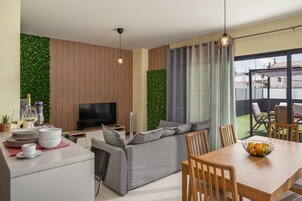 Una sala moderna con televisor de pantalla plana, sofá gris, mesa de comedor con sillas y un balcón con mobiliario exterior.