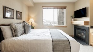 A bedroom with a large bed, a fireplace, a TV mounted on the wall, and a window with a view of the outside.