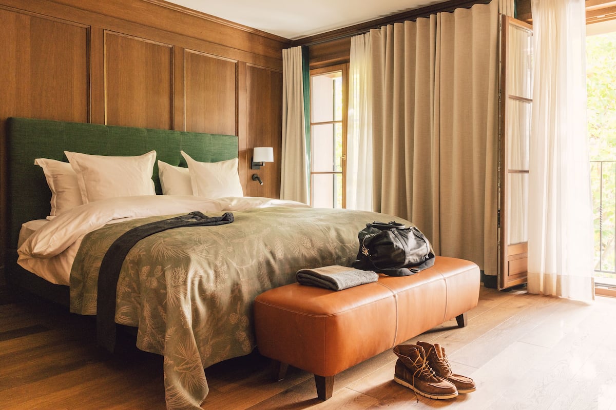 Una cama bien tendida con cabecera verde, un banco con un bolso y zapatos, y una ventana con cortinas.