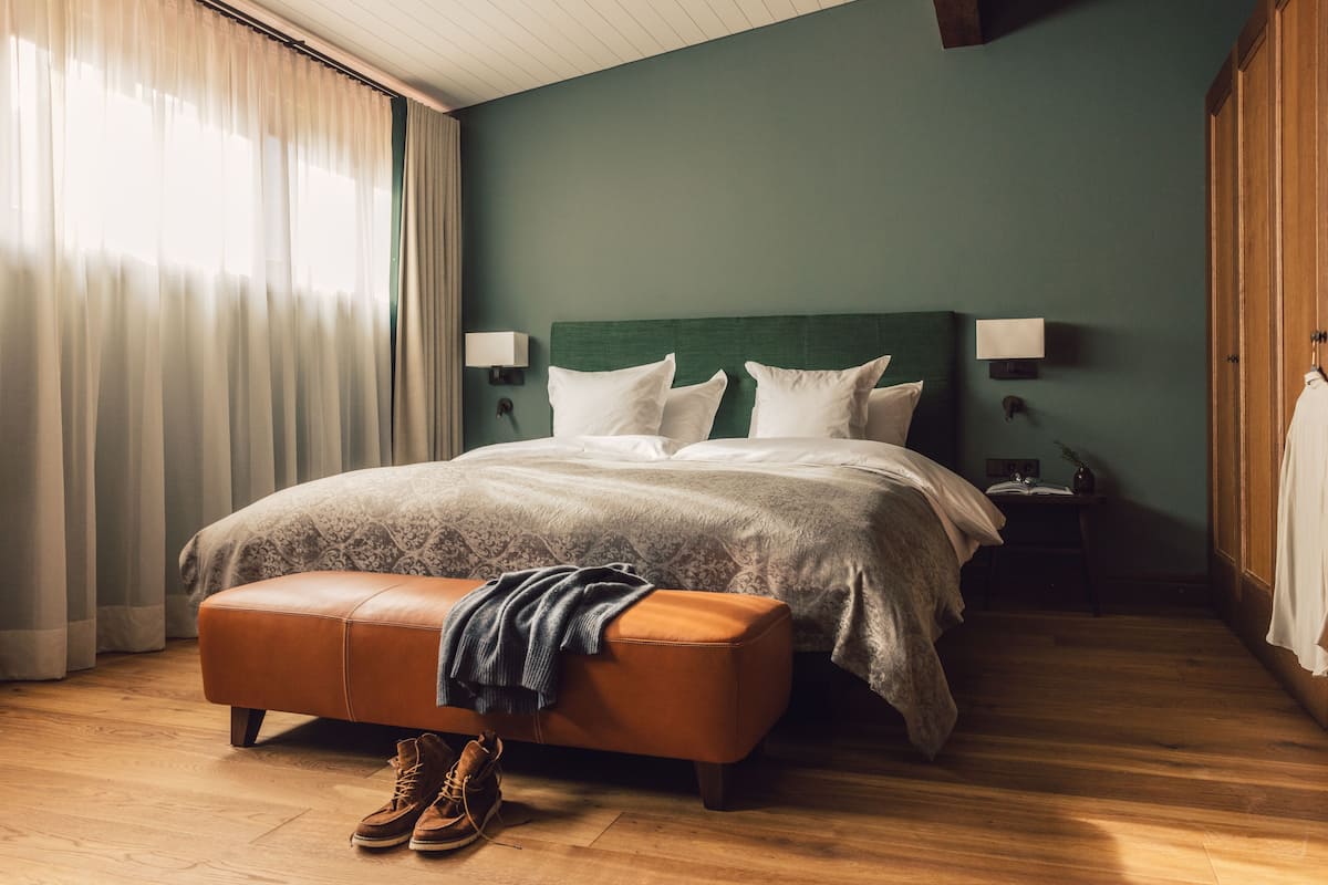 Una cama bien hecha con cabecera verde, un banco de madera con una manta, y un par de zapatos en el suelo.