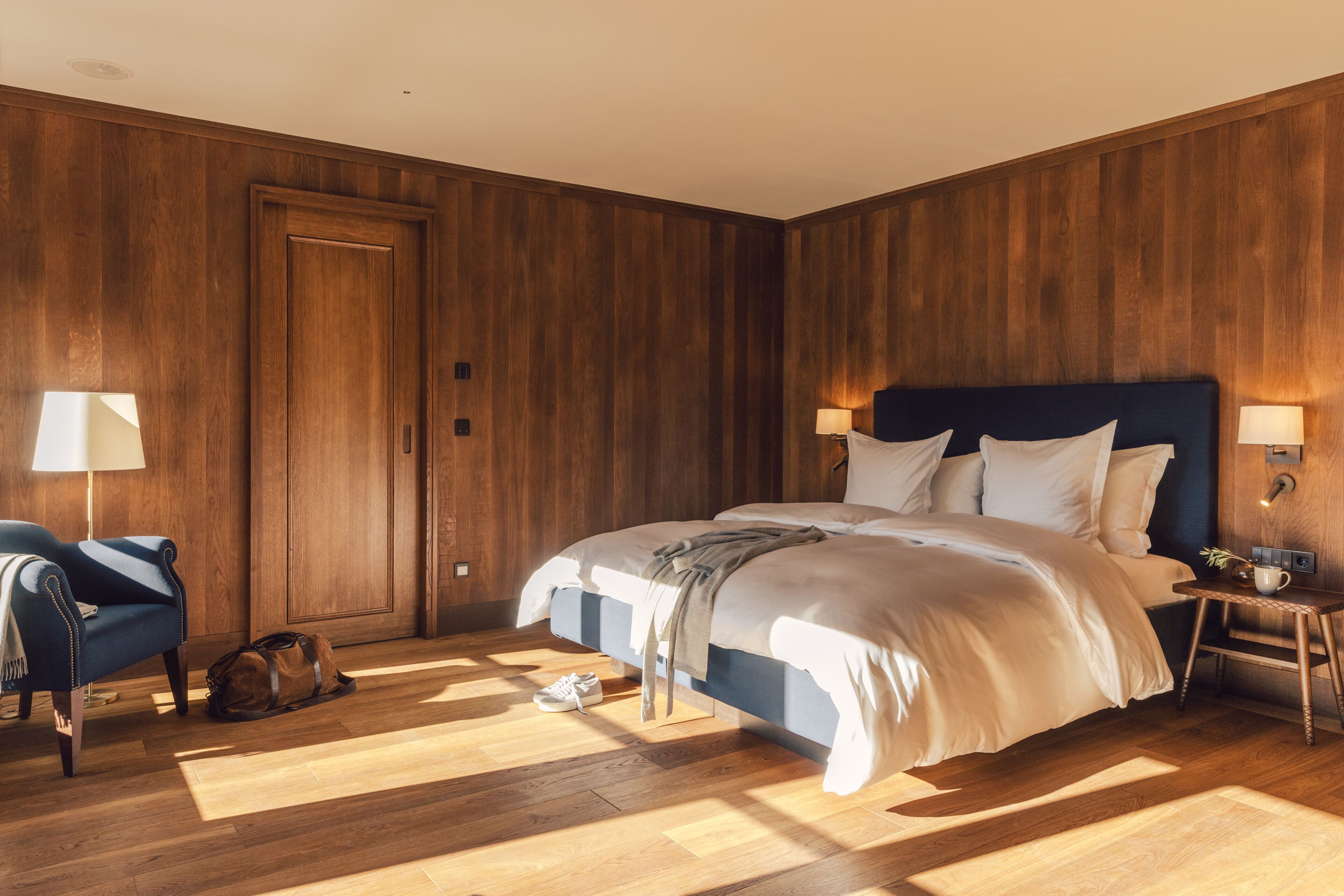 A bedroom with a large bed, wooden walls, a chair, and a nightstand with a lamp.