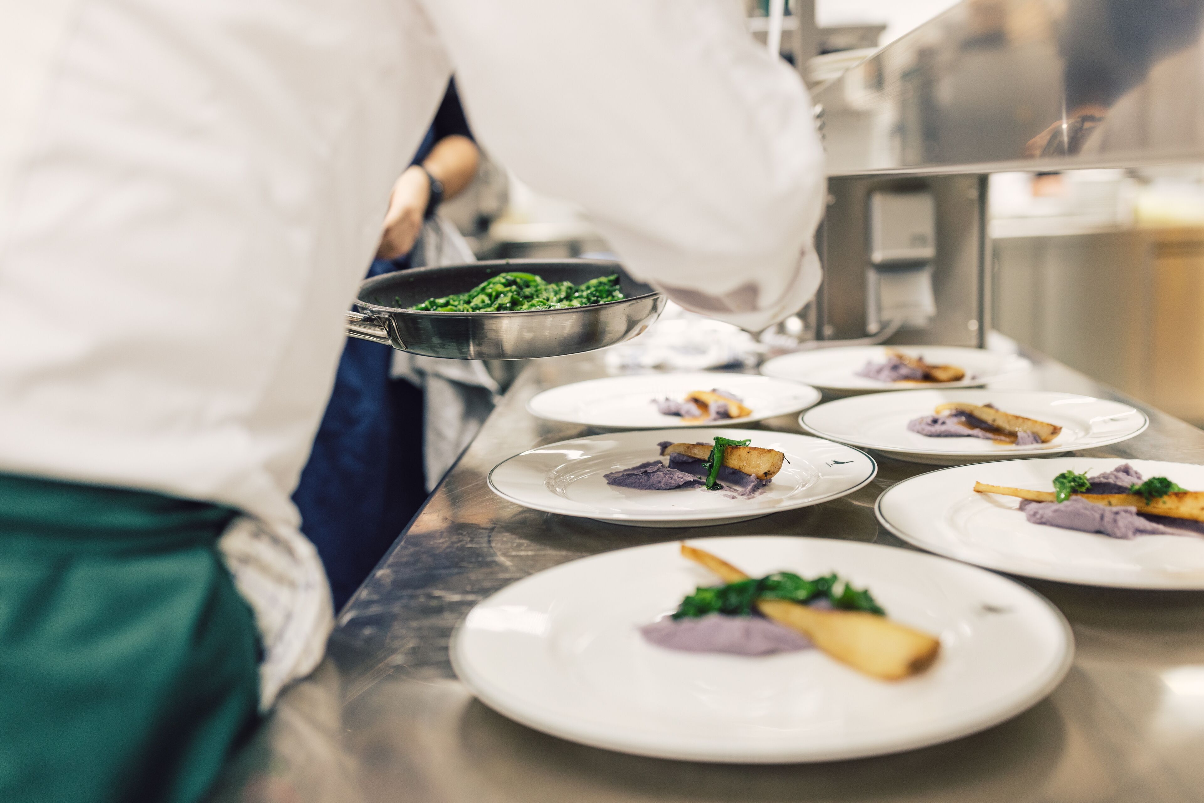 Una encimera de cocina con platos de comida, una persona con bata blanca y un bol de metal con verduras.
