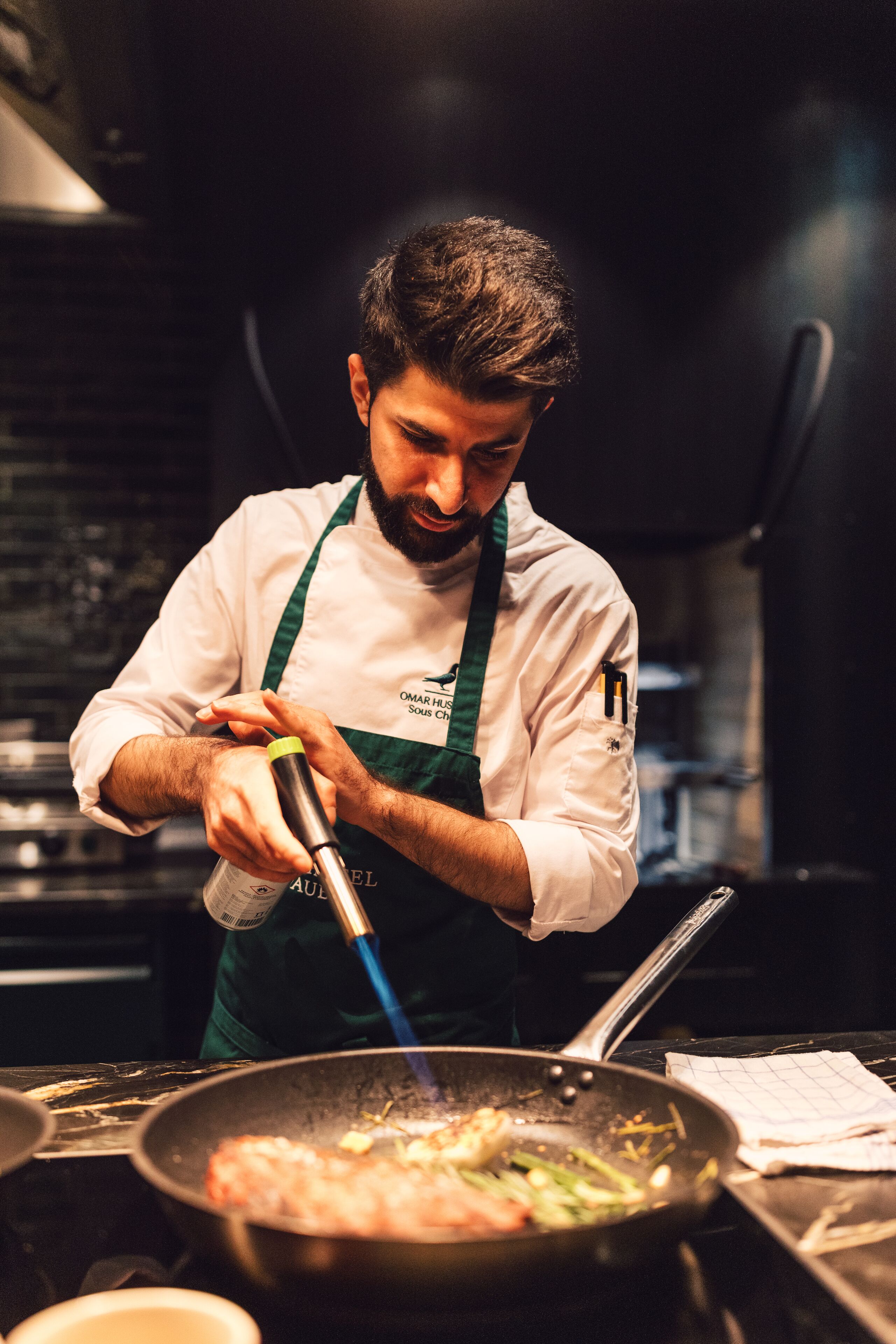 A chef in a kitchen, wearing a white chef's coat and a green apron, cooking with a pan on a stove.
