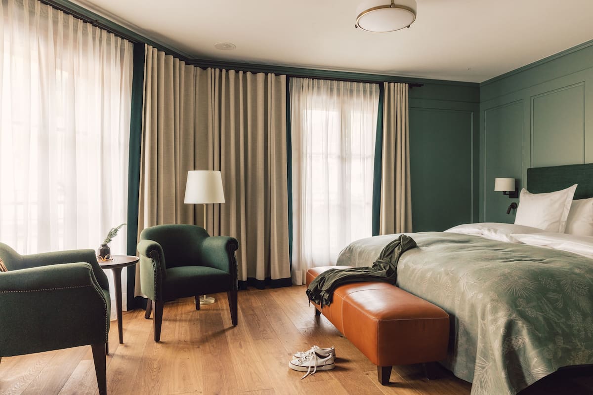 Una habitación de hotel con una cama, un sillón verde, una mesita de noche con una lámpara y un banco.