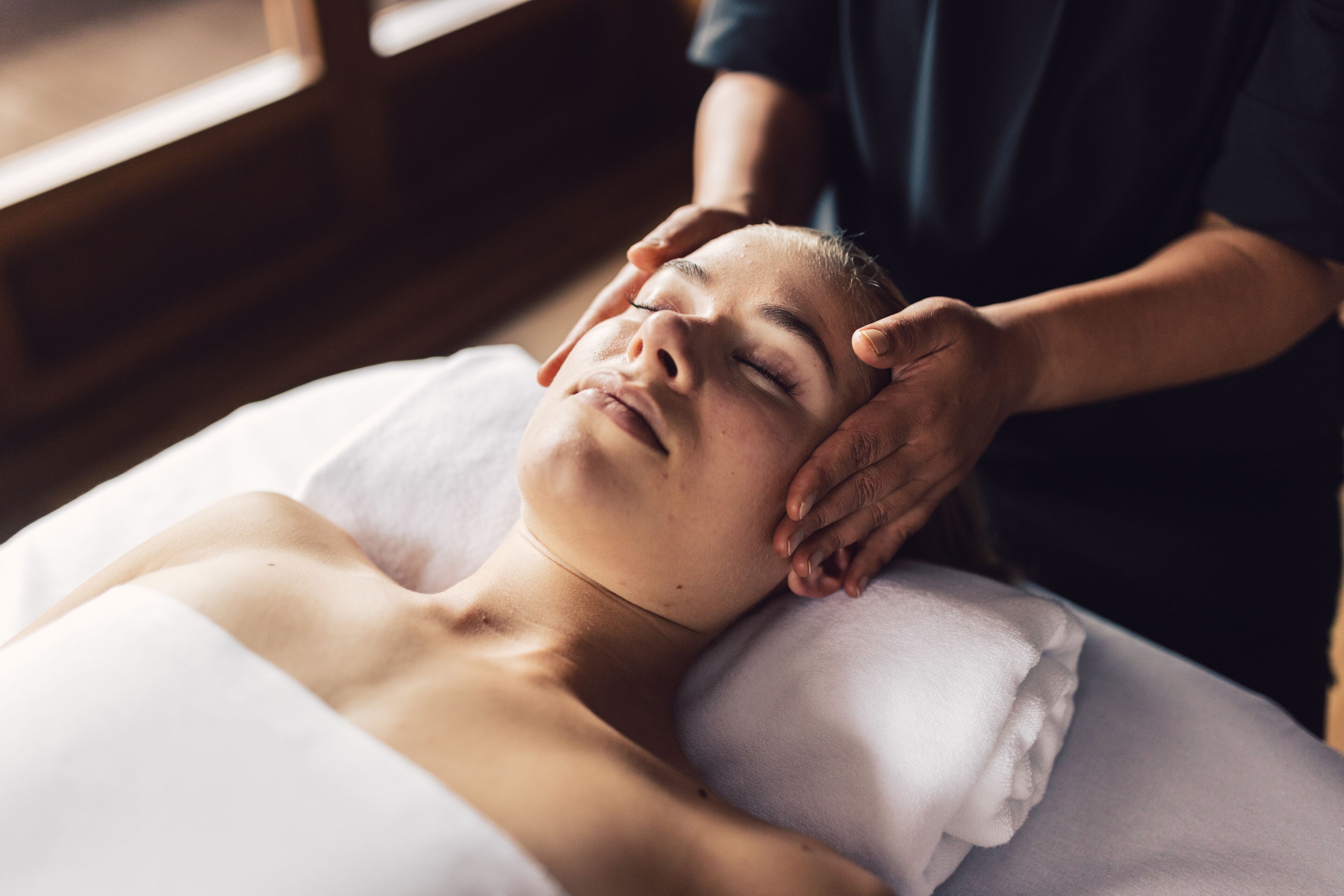 A person lying on a bed with a white pillow, receiving a head massage from another individual.