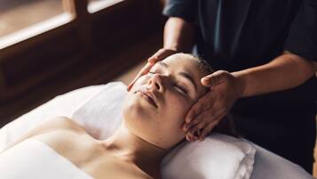 A person lying on a bed with a white pillow, receiving a head massage from another individual.