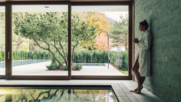 A person in a robe by a pool with a view of trees and a building. - Posthotel Taube (Schruns)