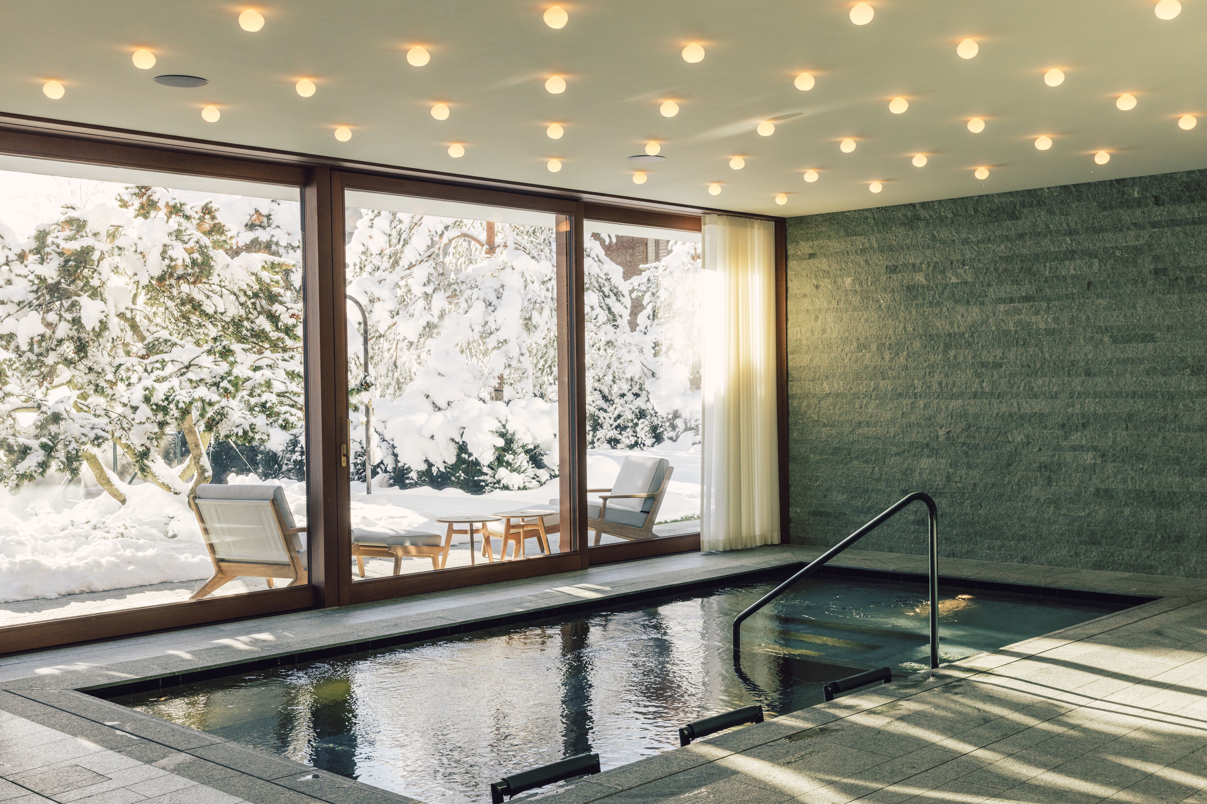 A modern indoor pool with clear glass walls, a wooden deck, and a stone wall.