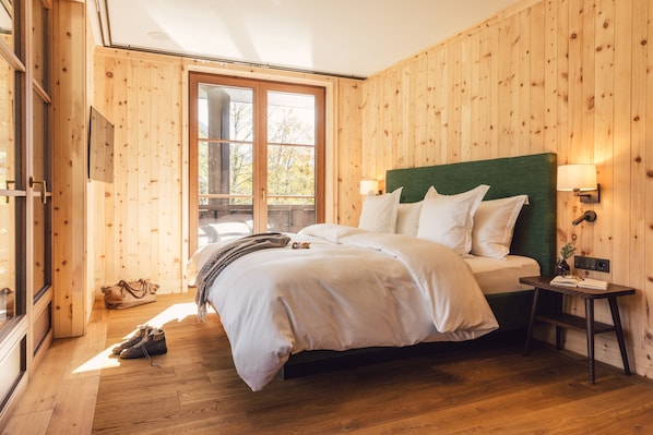 A bedroom with a wooden paneled wall, a large window, a bed with white bedding, a bedside table with a lamp, and a pair of shoes on the floor. - Posthotel Taube (Schruns)