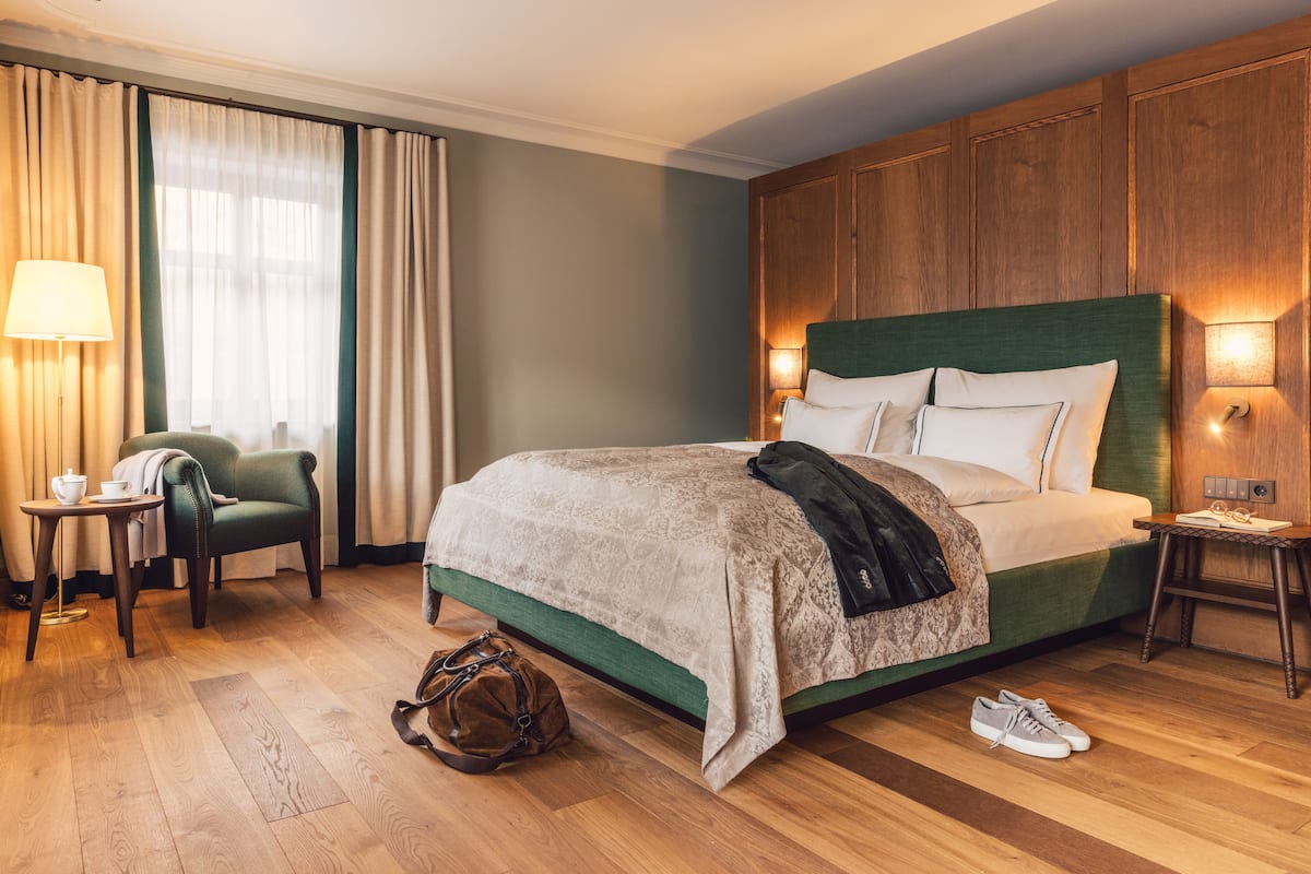 Habitación de hotel con una cama grande, una silla, una mesa con lámpara y una maleta en el suelo.
