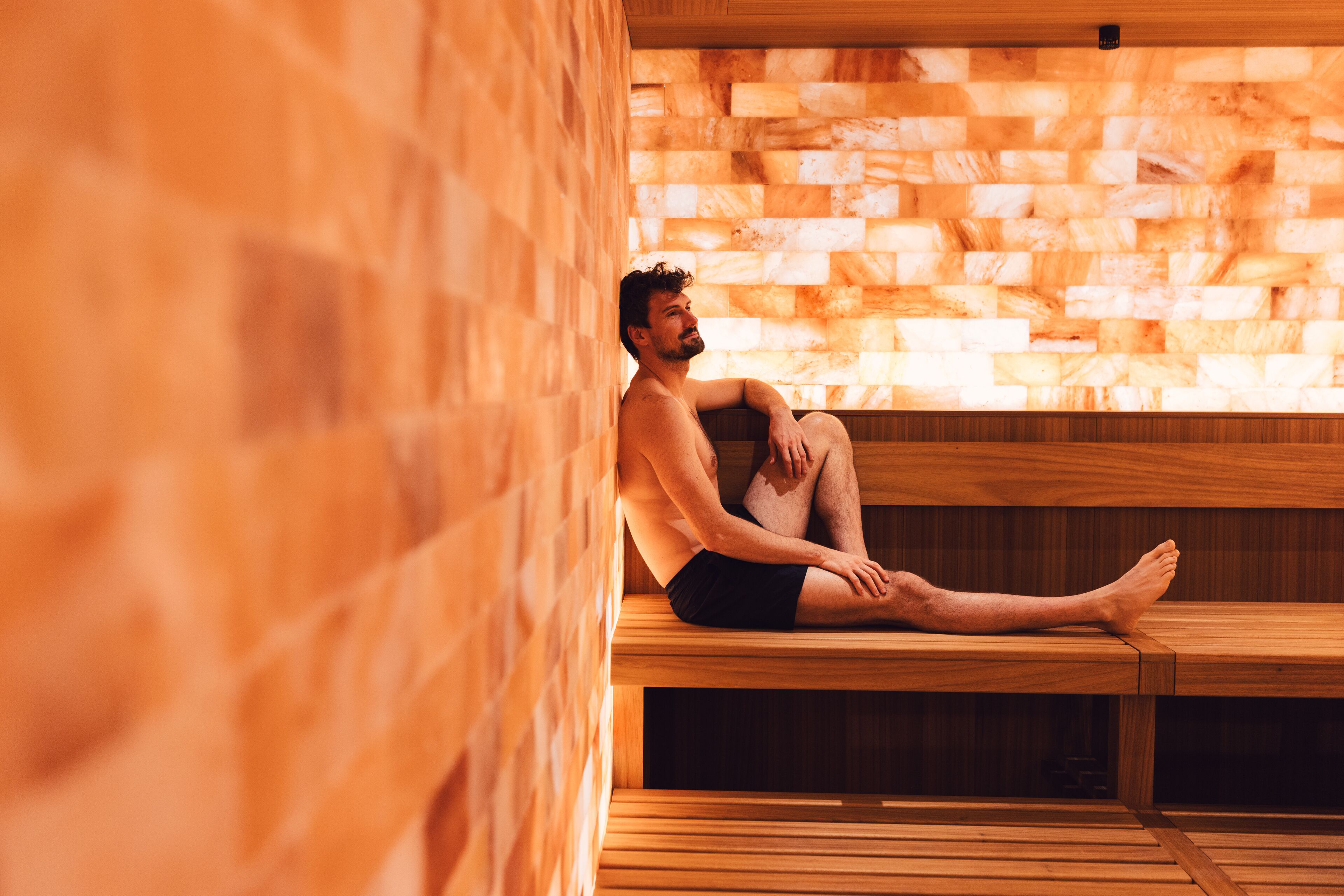 Eine Person sitzt auf einer Holzbank in einer Sauna mit einer Ziegelwand im Hintergrund.