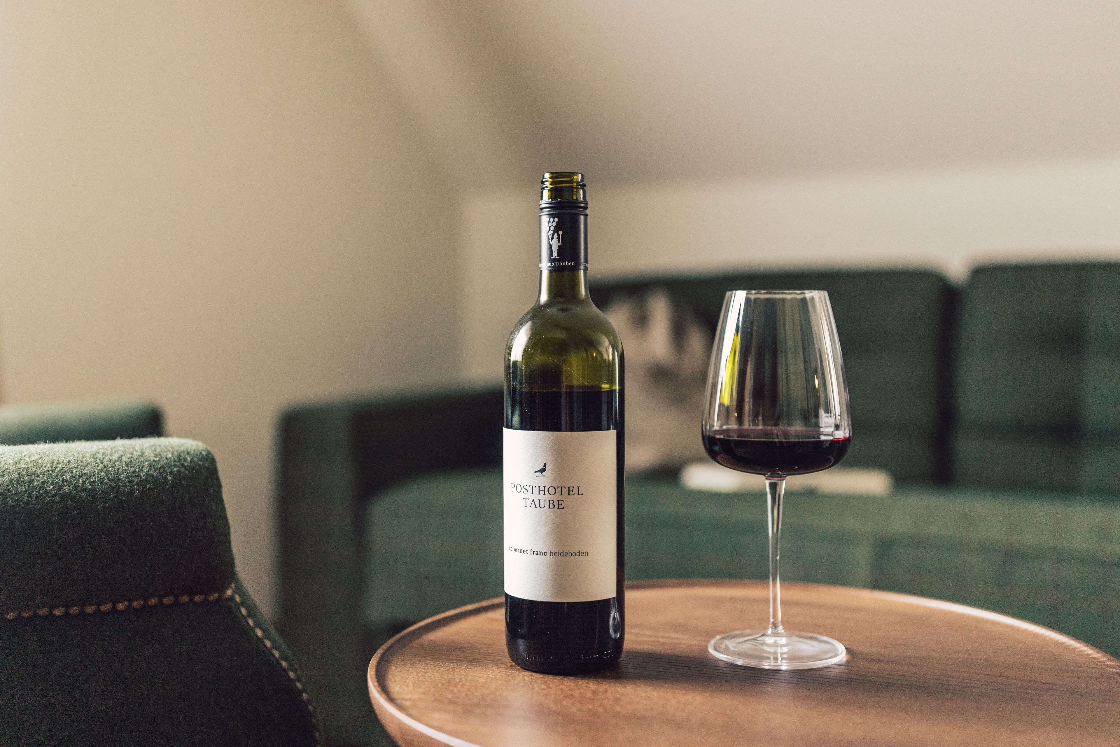 A bottle of wine and a glass on a wooden table in a room with green upholstered furniture.