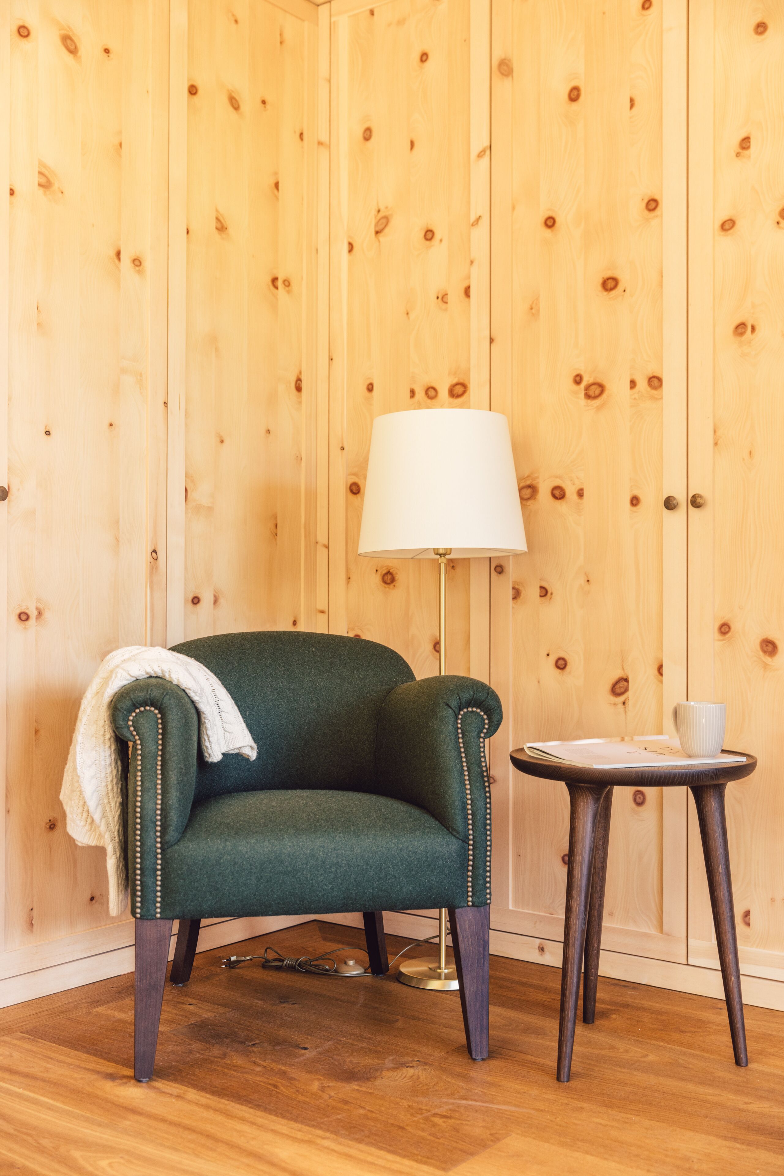 A room with a green armchair, a small round table with a lamp, and a wooden wall.