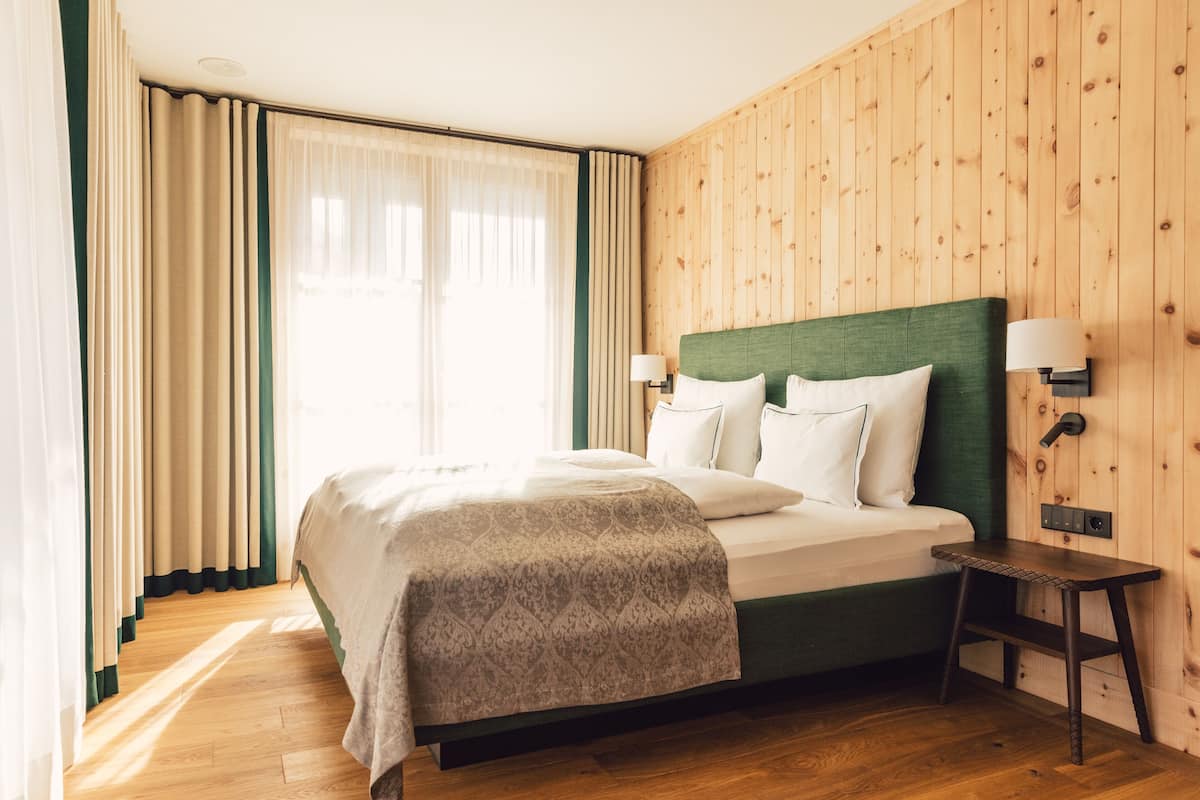 Una habitación de hotel con un revestimiento de madera, una cama grande con cabecera verde, dos lámparas de noche y una mesita de noche de madera.