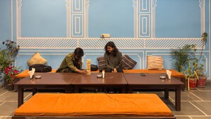A room with a blue wall featuring traditional patterns, a wooden table with orange cushions, and potted plants. - Madpackers Udaipur (Udaipur)