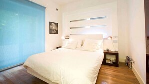 A modern bedroom with a large bed, a bedside table with a lamp, and a wall-mounted shelf. - Entire apartment in Ravda (Ravda, Nessebar community)