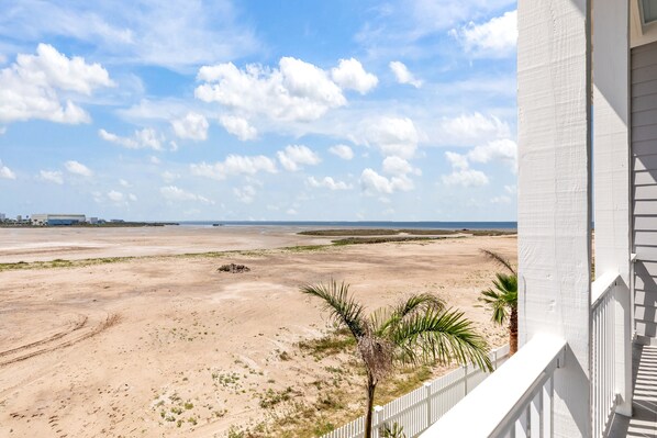 Vista a la playa desde un balcón con barandilla blanca, playa de arena y palmeras.