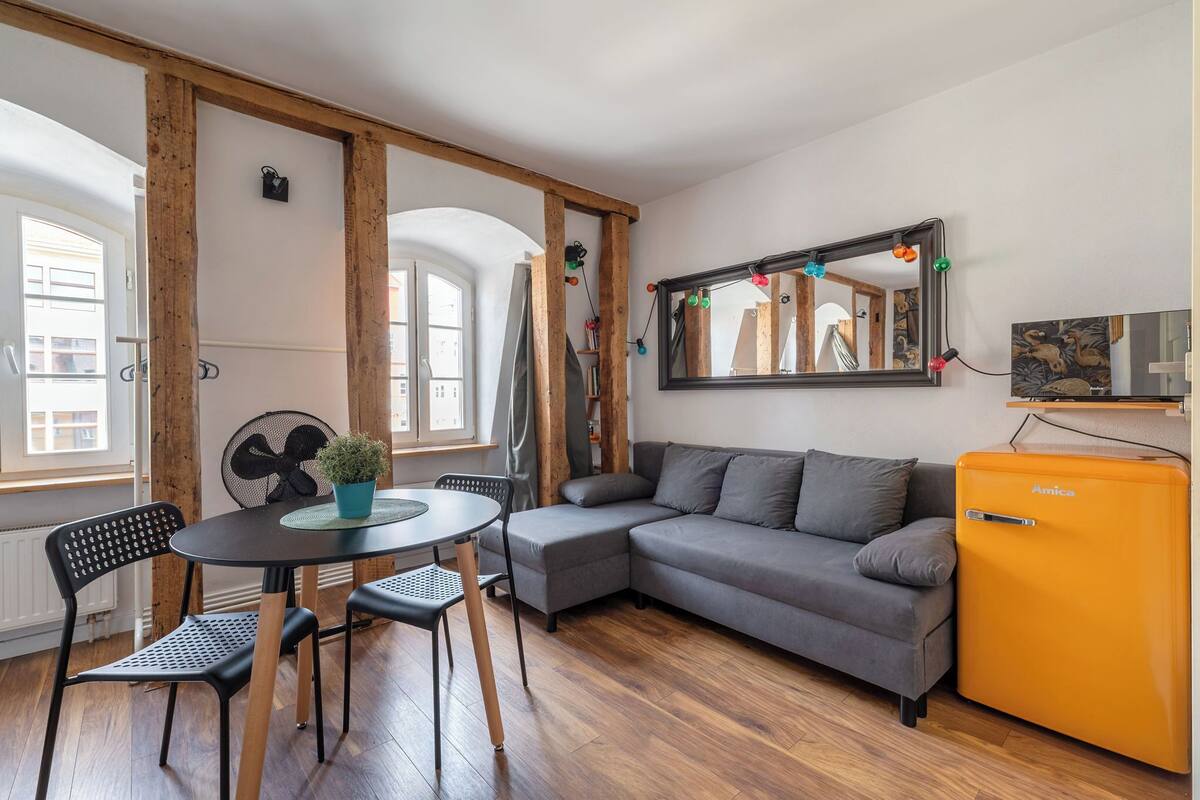 Una sala de estar acogedora con un sofá gris, un refrigerador naranja pequeño, una mesa redonda con sillas y un espejo grande en la pared.