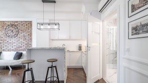 A modern kitchen with white cabinets, a brick accent wall, and a marble countertop. There are two stools and a hanging light fixture.
